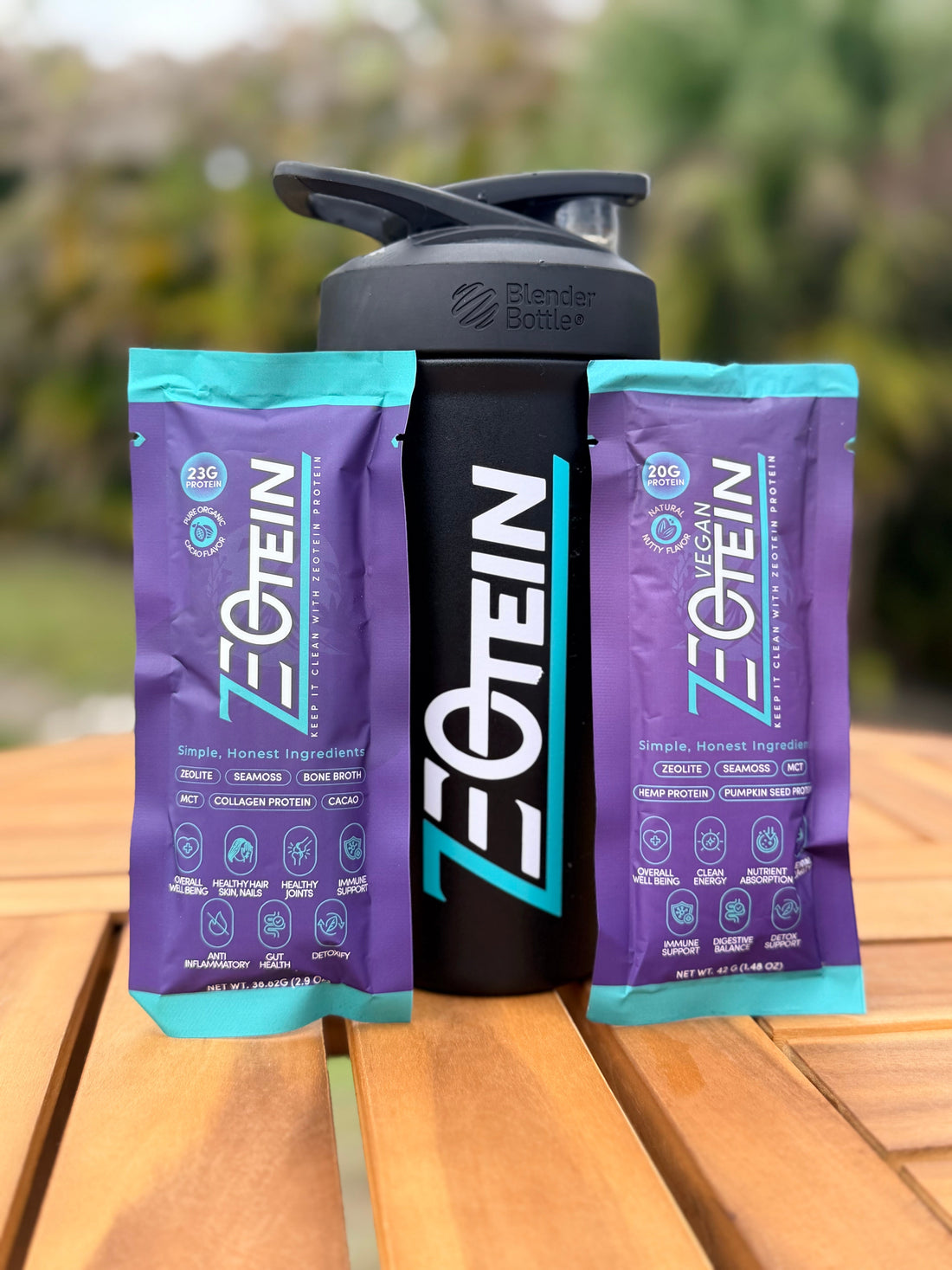 Two Zeotein single serve stick packs standing either side of a black Zeotein shaker bottle on a wooden table outdoors