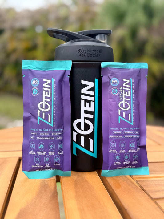 Two Zeotein single serve stick packs standing either side of a black Zeotein shaker bottle on a wooden table outdoors
