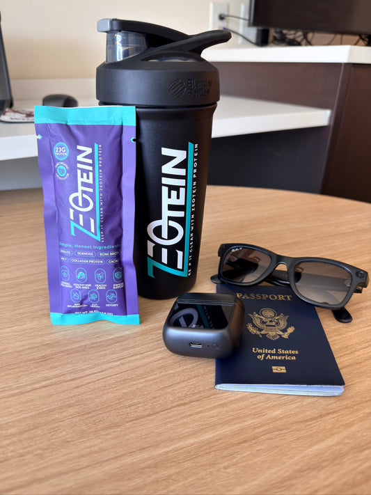 A Zeotein single serve stick pack and a black Zeotein shaker bottle on a desk with sunglasses, earbuds and a passport, packed for travel