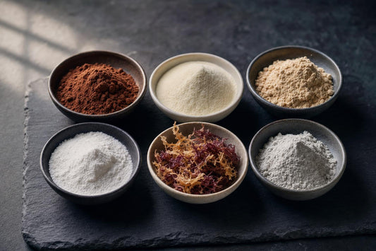 Six small ceramic bowls holding the ingredients in Zeotein Cacao: cacao powder, collagen peptides, bone broth protein, MCT powder, dried sea moss and zeolite mineral